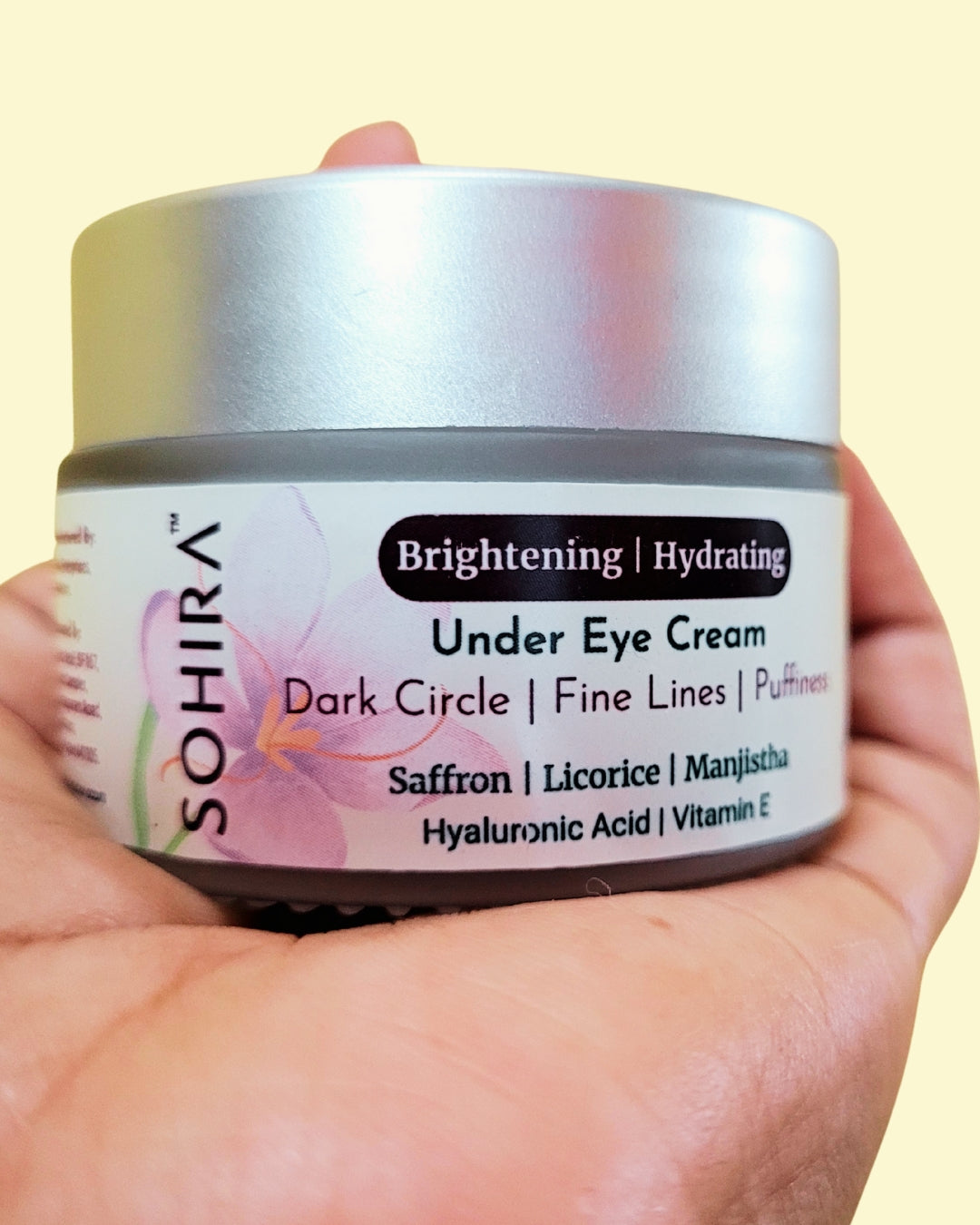 Hand holding a jar of SOHIRA Under Eye Cream against a light yellow background